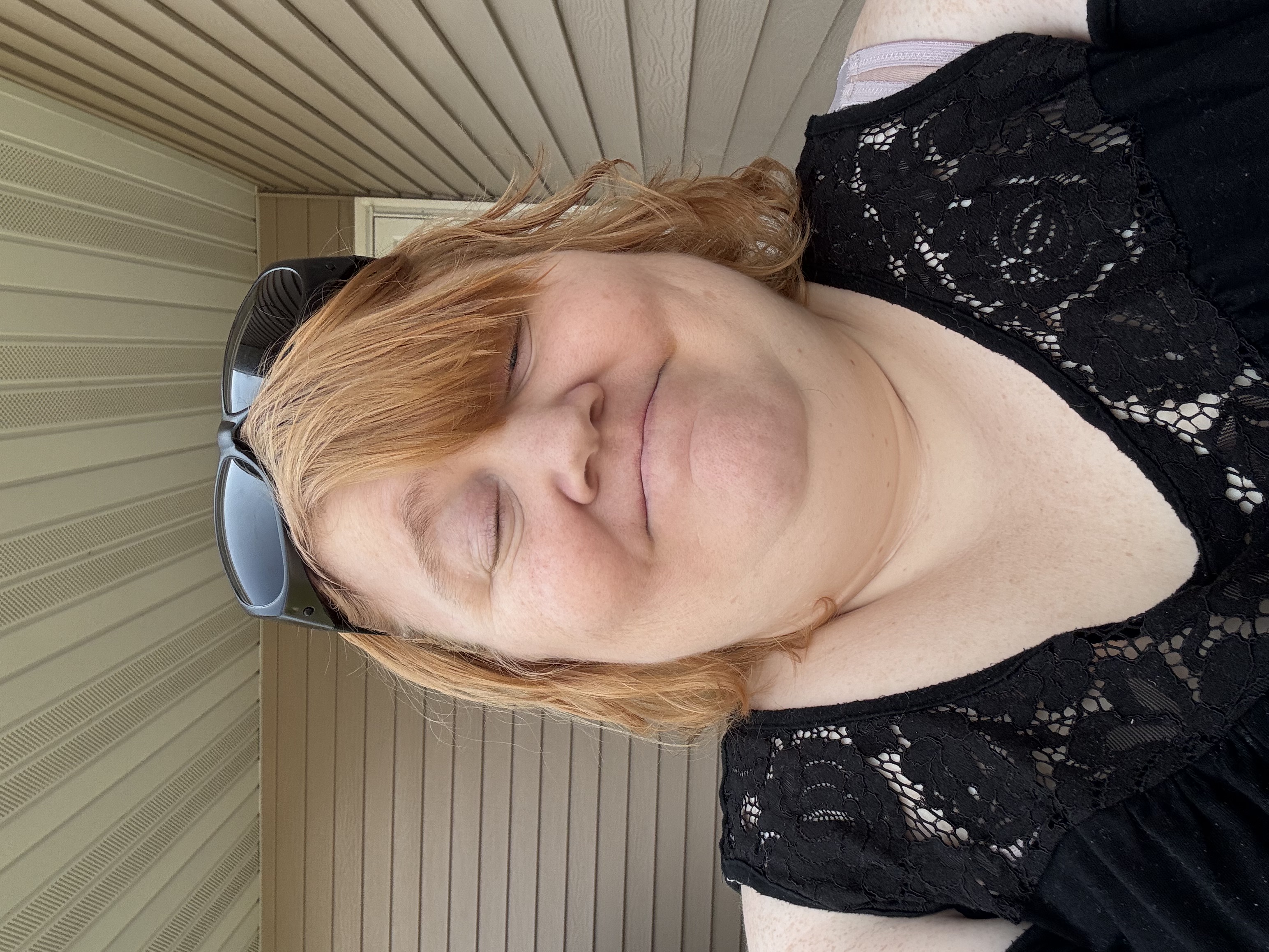 A picture of Sabrina Chavez, the Founder of Jessica & Sabrina's Crafty Creations: A close-up portrait of a smiling woman with red hair and sunglasses on her head, wearing a black lace top outdoors beneath a porch.
